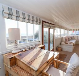 A room with a wooden desk, a lamp, a striped chair, a sectional sofa with cushions, and windows showing a beach view at Valetta, Thorpeness in Thorpeness