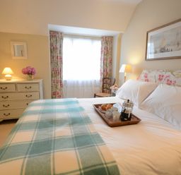 A bedroom with a bed covered with a check blanket and a tray with coffee and croissants on it a chest of drawers with a lamp and flowers near a window with floral curtains at 2 The Bays Thorpeness