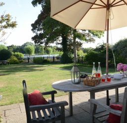 An outdoor patio with a wooden table and chairs under an umbrella overlooking a garden with a lake in the background at 2 The Bays Thorpeness