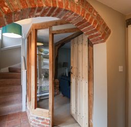 A hallway with a brick arch, wooden door, glass panels, staircase with carpet, and a blue lamp shade at 1 The Old Coach House Huntingfield Near Laxfield