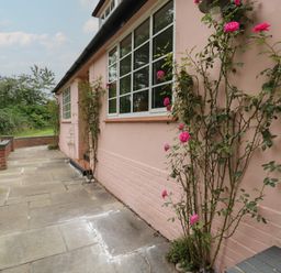 A paved walkway along a pink exterior wall with climbing rose plants and a brick low wall at Ash Tree Cottage in Aldringham