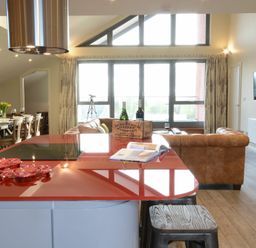An open plan living area with a red kitchen island, brown sofas, large windows, and a dining table with chairs at King George Penthouse in Aldeburgh