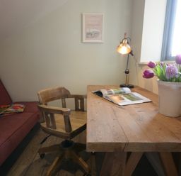 A wooden desk with a chair lamp an open book and flowers next to a window and a red sofa with books in a room at King George Penthouse in Aldeburgh