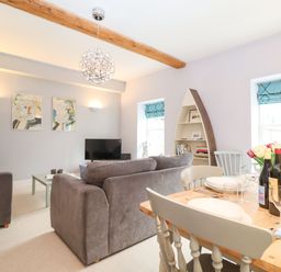 A living room with gray sofas a coffee table a TV a boat-shaped bookshelf and a dining table with wine bottles and flowers at Saffron Blythview in Blythburgh near Southwold
