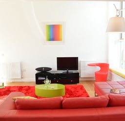 A living room with a red sofa a green coffee table a red rug and a TV on a black stand with a rainbow artwork on the wall at Parrotfish Bay in Southwold