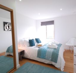 A bedroom with a double bed blue pillows and blankets a window with a blind a white chair a dresser and a mirror at Seal Cove in Southwold