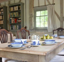 A dining table set with blue and white china teacups and bowls a silver teapot and a plate of pears in a room with wooden beams bookshelves and a window at The Gig House Stowupland