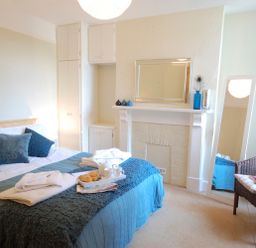 A bedroom with a bed covered with blue and gray bedding and pillows a tray with croissants and cups on the bed a white fireplace with a mirror and decorative items a wicker chair with cushions and a tall mirror near the window at Spindrift Southwold