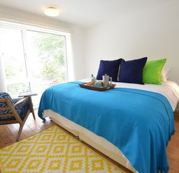 A bedroom with a bed covered in a blue blanket and pillows a blue patterned chair a yellow rug and a small white nightstand with a blue lamp at Aldeburgh Lodge Gardens in Aldeburgh