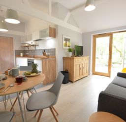 A kitchen and dining area with a round wooden table set with dishes and drinks a wooden sideboard with a TV a gray sofa and glass doors opening to a garden at Milk Parlour in Spexhall Near Halesworth