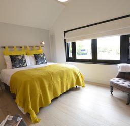 A bedroom with a double bed yellow blanket and pillows a gray chair by the window and a small wooden side table at Milk Parlour Spexhall Near Halesworth