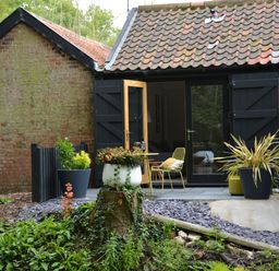 A brick building with black wooden shutters and a tiled roof with potted plants and outdoor chairs at Milk Parlour Spexhall Near Halesworth
