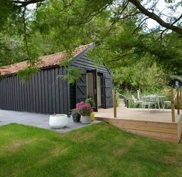 A black wooden barn with open doors next to a wooden deck with a white table and chairs in a garden at Red Poll Barn Spexhall Near Halesworth