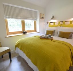 A bedroom with a bed covered in a yellow blanket and matching pillows next to a window and a chair at Red Poll Barn in Spexhall Near Halesworth