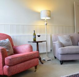 A sitting area with a red armchair and a beige loveseat with cushions a wooden side table with a wine bottle and glass and a floor lamp at Walton House Southwold
