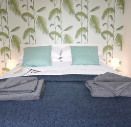 A bed with folded towels on a blanket and pillows against a wall with palm leaf wallpaper at Sweet Pea Southwold