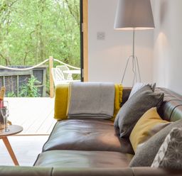 A brown leather sofa with cushions and a throw next to a standing lamp and a small table with a bottle and glasses near an open door showing a deck and greenery outside at Spexhall Barns in Halesworth