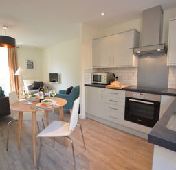 A kitchen and dining area with a round wooden table set for two and a living room with chairs and a small tv at Mycroft Southwold in Southwold