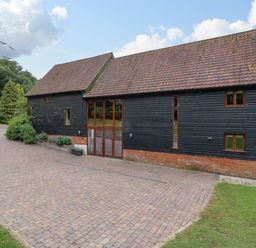 Exterior view of a barn style building with black wooden siding and tiled roof next to a paved driveway and green lawn at Quill Farm Barn in Campsea Ashe