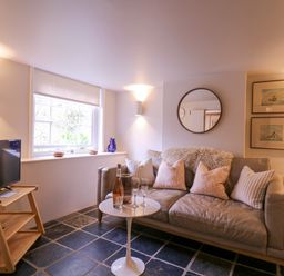 A living room with a gray sofa and pillows a small round table with bottles and glasses a television on a wooden stand and framed pictures on the wall at Mariners Cottage King Street in Aldeburgh