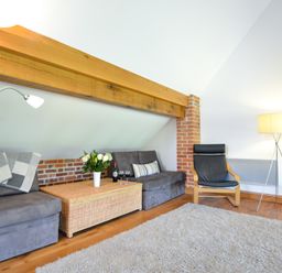 A living room with a gray sofa a wicker coffee table with flowers and wine a black armchair and lamps at Two Chantry Barns Orford