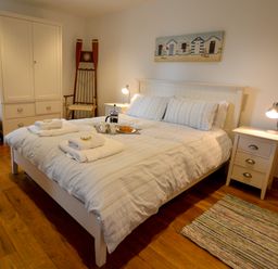 A bedroom with a double bed and striped bedding two bedside tables with lamps a wicker basket a wooden chair a white wardrobe and a small rug on wooden floor at Two Chantry Barns Orford