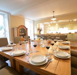 A dining room with a large wooden table set with plates glasses and cutlery and a kitchen with beige cabinets in the background at Henstead Hall in Henstead near Beccles