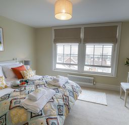 A bedroom with a bed with folded towels pillows and a tray a window with blinds a lamp a painting on the wall and a dressing table with stool and flowers at 10 Church Walk Aldeburgh