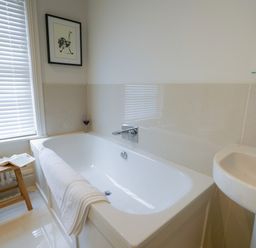 A bathroom with a bathtub, a white towel on the bathtub, a wooden stool with a flower vase and a book, a sink, and a window with blinds at 10 Church Walk, Aldeburgh in Aldeburgh