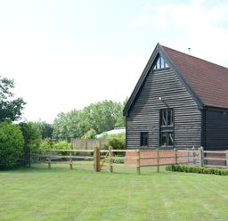 A wooden barn with a red roof next to a fenced lawn and trees at Manor House Barn Peasenhall