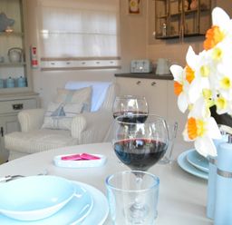 A dining table with plates wine glasses and flowers in a room with an armchair and kitchen cabinets at The Nookery at Snape Hall in Snape Suffolk