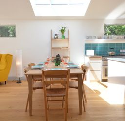 A dining area with a wooden table set with plates and glasses next to a kitchen with teal tiles and a yellow armchair in 2 Pink Cottages Aldeburgh
