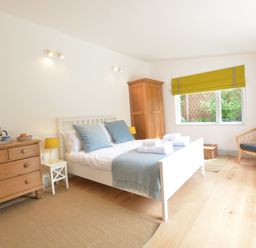 A bedroom with a white bed and blue pillows next to a wooden dresser and a wooden chair at 2 Pink Cottages Aldeburgh