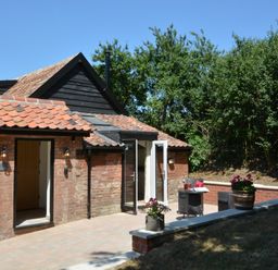 A brick house with tiled roof and open doors leading to a patio with a small table and two chairs with flowers on the wall at Carriage House Bruisyard Near Framlingham
