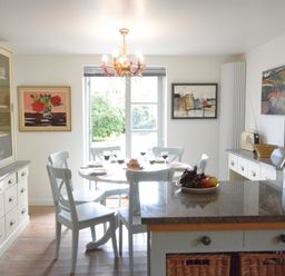 A dining room with a round table set with plates and wine glasses surrounded by chairs and a kitchen island with a bowl of fruit at Robert Sadler's Studio in Aldeburgh