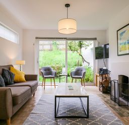 A living room with a sofa with yellow and dark pillows two gray chairs a coffee table a fireplace and a wall-mounted TV at The Bridge House in Southwold