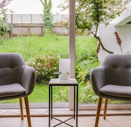 A sitting area with two grey chairs and a small table with a white lamp in front of a glass window showing a garden at The Bridge House Southwold
