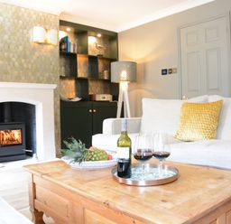 A living room with a wooden coffee table holding a wine bottle and glasses in front of a white sofa with a yellow pillow at The Red Brick House Aldeburgh