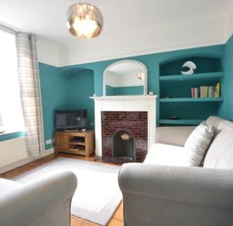 A living room with teal walls a fireplace a mirror a television on a wooden cabinet shelves with books and decorative items grey sofas and a window with patterned curtains at Somewhere Only We Know in Aldeburgh