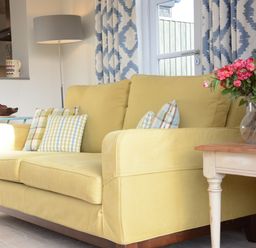 A yellow sofa with patterned cushions next to a small table with a vase of flowers and a floor lamp behind at Mellows Thorpeness in Thorpeness