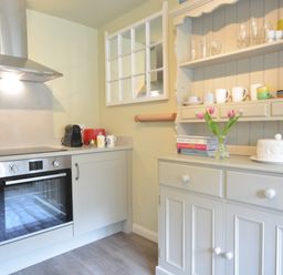 A kitchen with an oven and stove next to a cupboard with shelves holding cups and glasses with flowers on the countertop at Church Farm Cottage Dunwich Near Westleton