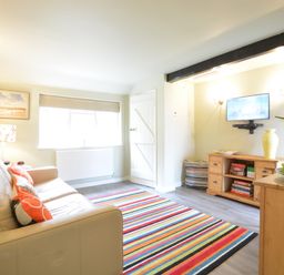 A living room with a beige sofa and orange pillows a striped rug a wooden cabinet with books and a yellow vase a wall-mounted television and a window at Church Farm Cottage Dunwich Near Westleton