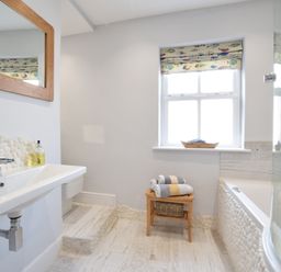 A bathroom with a sink mirror a small wooden stool with folded towels a window and a bathtub at 53 Tern Cottage Aldeburgh