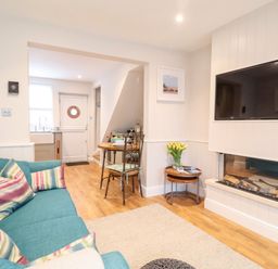 A living room with a teal sofa and colorful pillows a wall mounted TV and fireplace a small dining table and kitchen visible at 51 Tern Cottage Aldeburgh