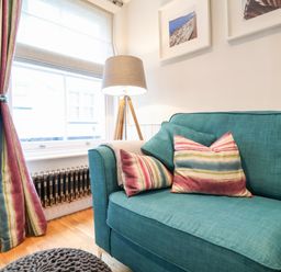 A corner of a living room with a teal sofa with striped cushions a window with striped curtains a floor lamp and framed pictures on the wall at 51 Tern Cottage Aldeburgh