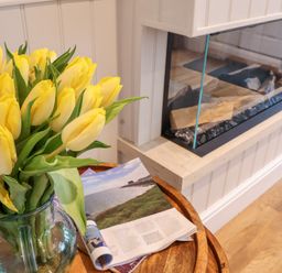 A bouquet of yellow tulips in a glass vase on a wooden table next to a magazine by a glass-enclosed fireplace at 51 Tern Cottage Aldeburgh