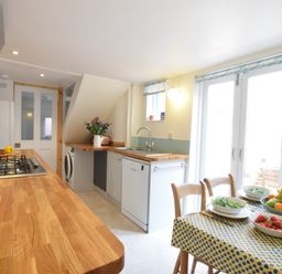 A kitchen with wooden countertops a gas stove a dishwasher washing machine a dining table with bowls of fruit and salad and double doors at Seaflower Aldeburgh