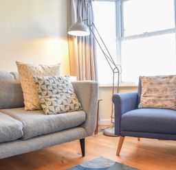 A living room with a grey sofa and a blue armchair with cushions and a floor lamp by the window at Seaflower in Aldeburgh