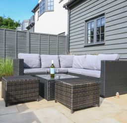 An outdoor patio with a gray cushioned sectional sofa and wicker furniture including a glass top table and two ottomans at Corner Cottage Thorpeness