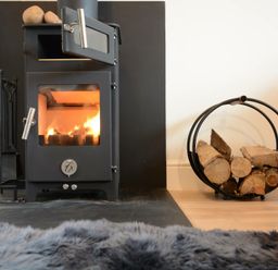 A wood burning stove with fire inside and firewood in a circular holder beside it at Fishpond House Sotherton in Halesworth